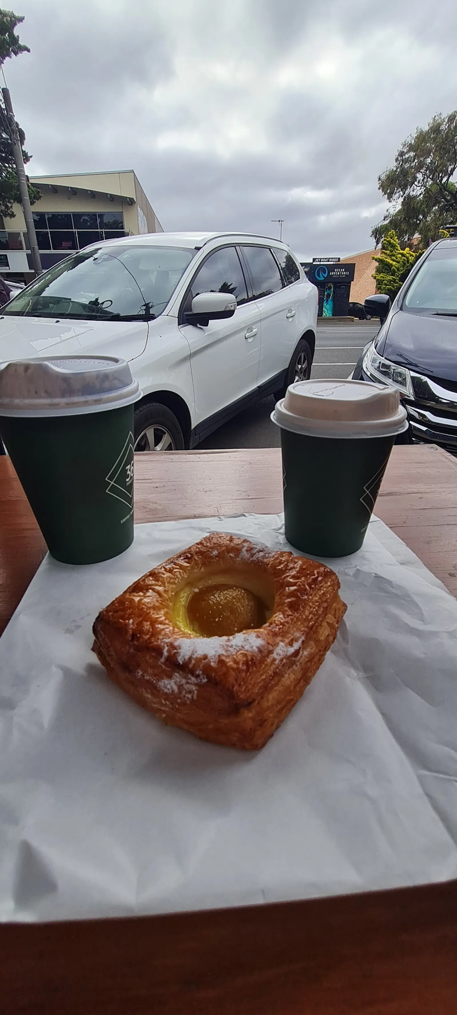 Two green takeaway coffees and a custard danish pastry on outdoor table at Cafe Biscotti