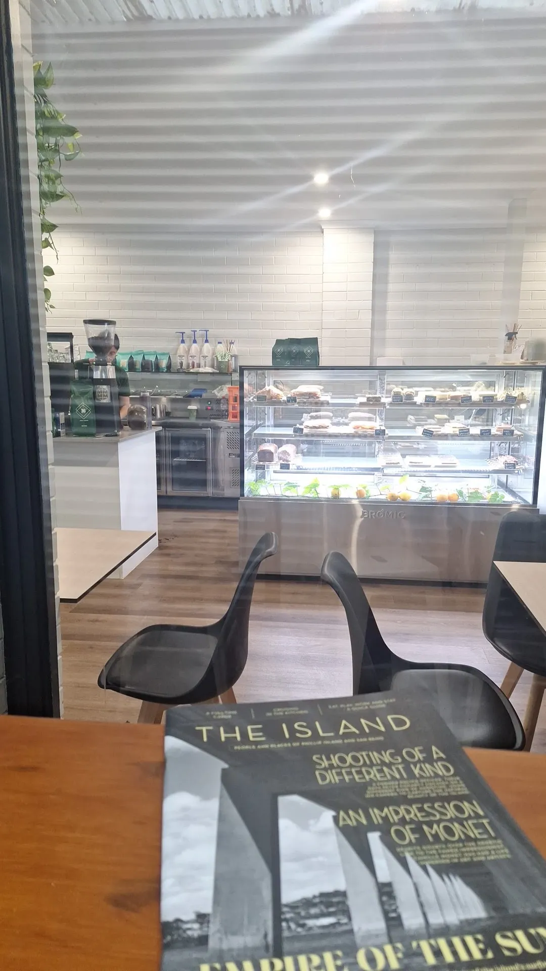 View through Cafe Biscotti window showing pastry display cabinet, Scandi chairs, and The Island magazine