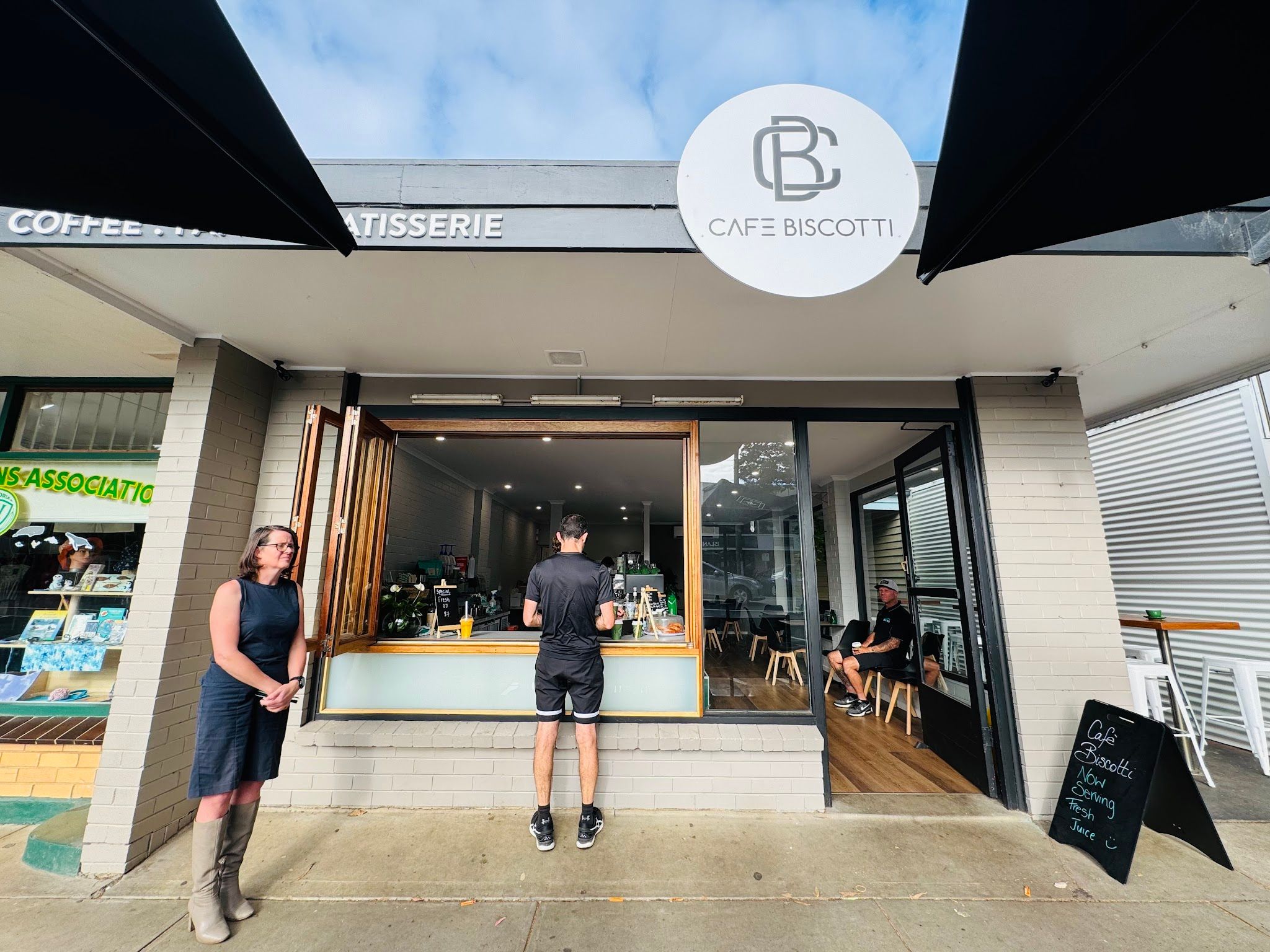 Cafe Biscotti storefront with barista at open window, CB logo above, woman standing outside Cowes